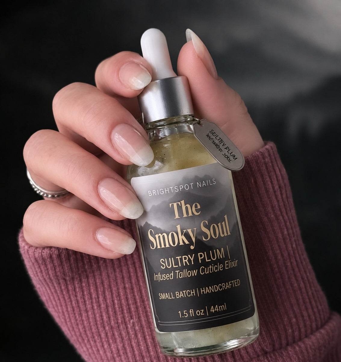 A hand with manicured nails holds a glass bottle of Brightspot Nails 'The Smoky Soul' Sultry Plum cuticle oil. The hand is wearing a dusty rose knit sweater, and the background is a dark, out-of-focus gray.